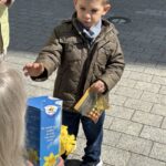 Boy in a brown quilted jacket extends his hand toward an older person outdoors on a paved sidewalk, with a blue donation box and yellow flowers nearby.