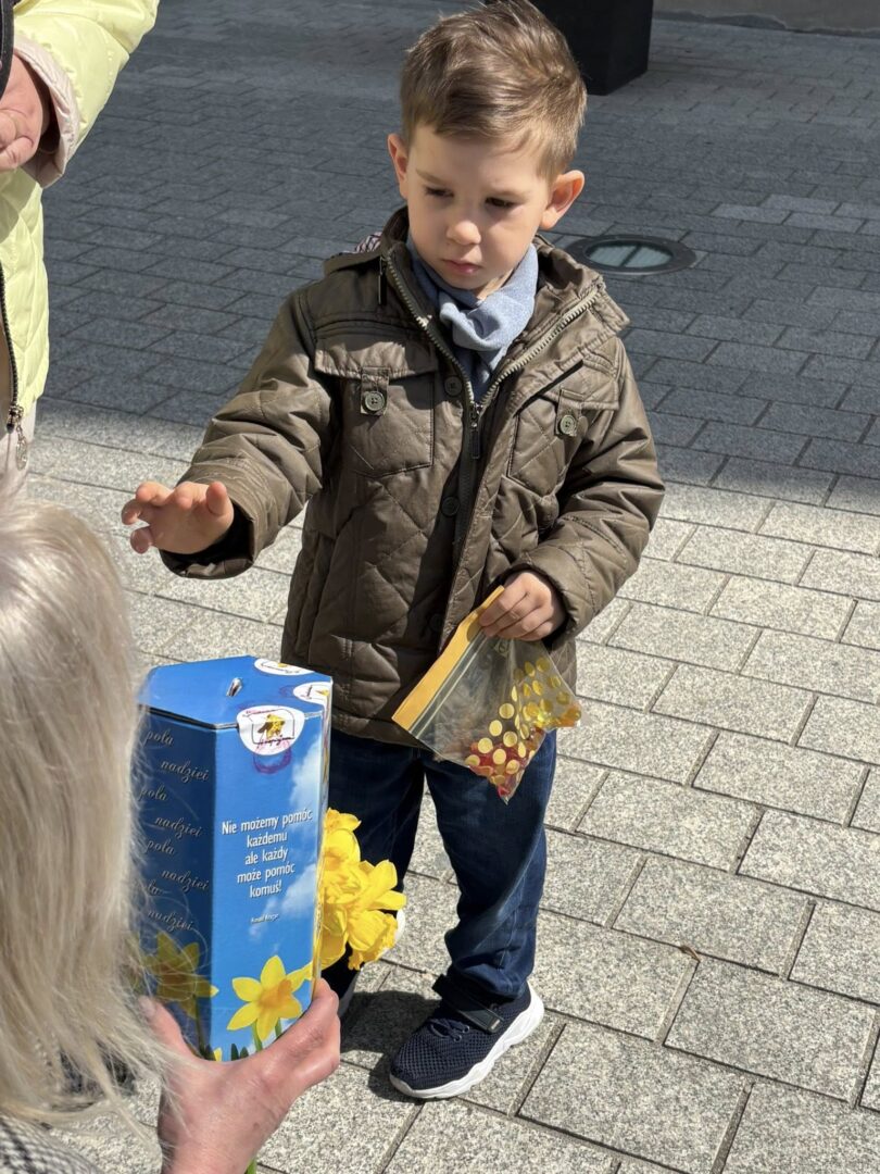 Boy in a brown quilted jacket extends his hand toward an older person outdoors on a paved sidewalk, with a blue donation box and yellow flowers nearby.