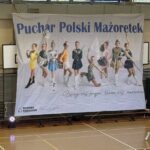 Banner for the Puchar Polski Mażoretek (Polish Majorette Cup) with a line of young baton twirlers in colorful uniforms posing in a gym hall.