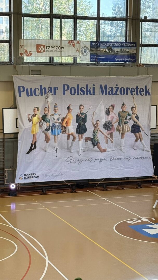 Banner for the Puchar Polski Mażoretek (Polish Majorette Cup) with a line of young baton twirlers in colorful uniforms posing in a gym hall.