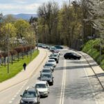 Queue of cars in traffic along a curved, tree-lined street on a sunny spring day; a pedestrian walks the left sidewalk.