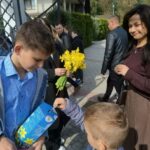 Older boy in a blue shirt hands a gift bag to a younger boy as a bouquet of yellow daffodils sits nearby at an outdoor event.