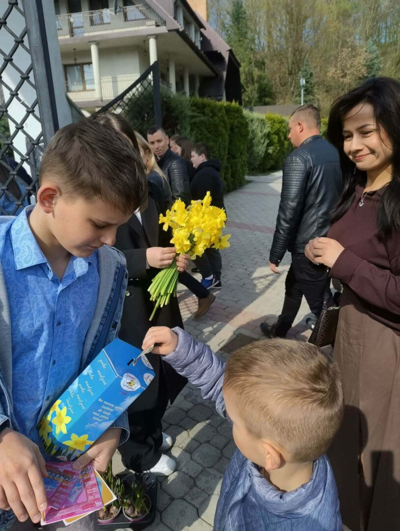 Older boy in a blue shirt hands a gift bag to a younger boy as a bouquet of yellow daffodils sits nearby at an outdoor event.