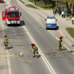 Emergency responders in protective gear sweep debris from a multi-lane road while a fire truck and police van stand by; pedestrians observe from the sidewalk.