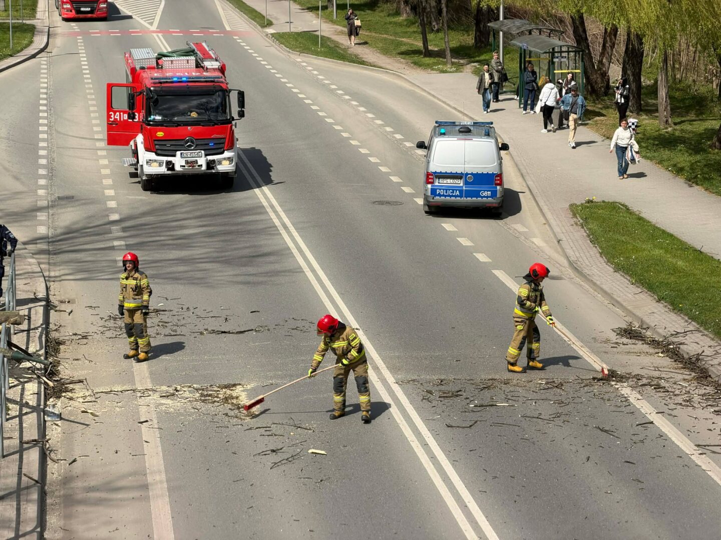 Emergency responders in protective gear sweep debris from a multi-lane road while a fire truck and police van stand by; pedestrians observe from the sidewalk.