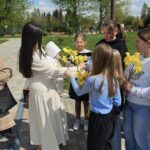Woman in a white dress hands yellow daffodils to a group of smiling children in a sunny park.