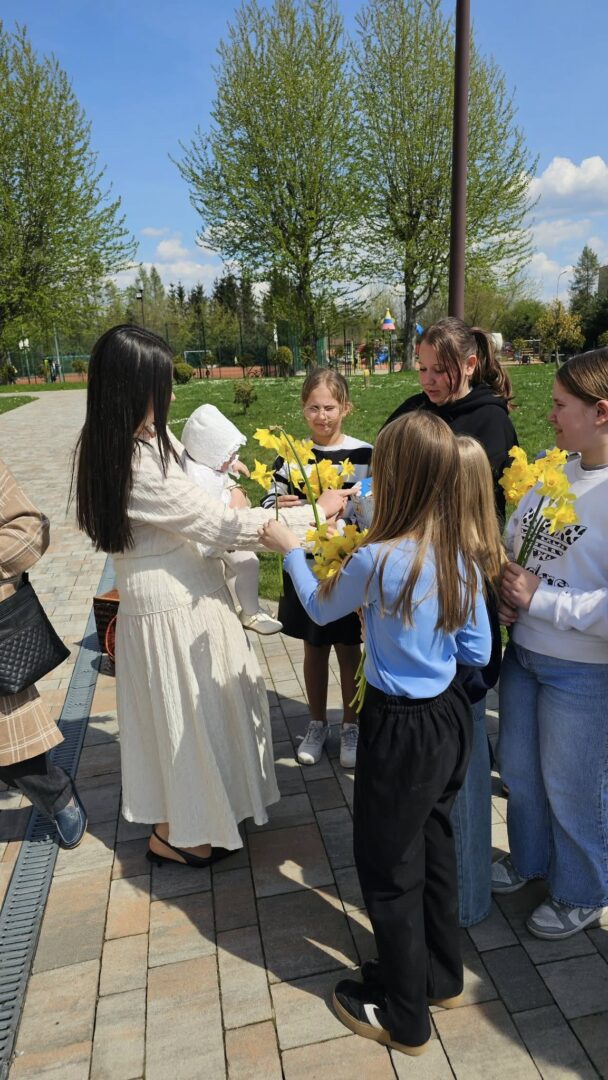 Woman in a white dress hands yellow daffodils to a group of smiling children in a sunny park.