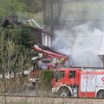 Residential area fire: firefighters on a roof amid smoke, with red fire trucks and a ladder extending toward the house