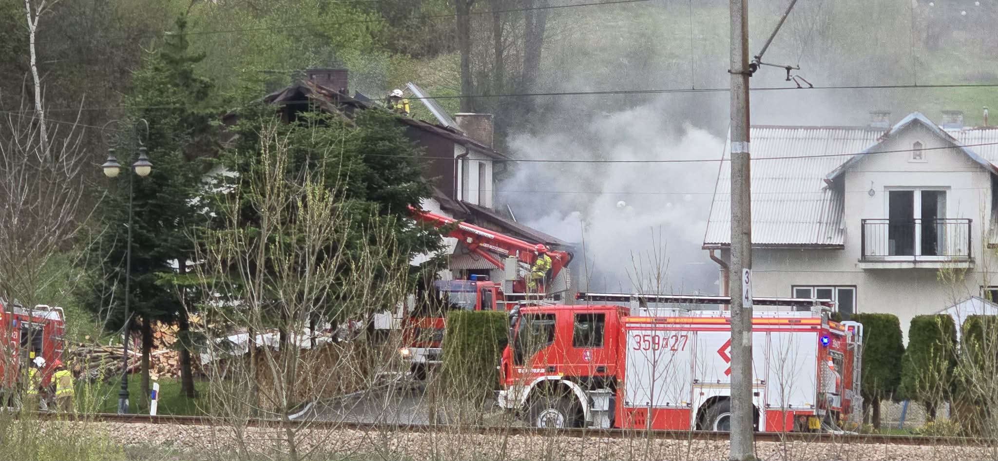 Residential area fire: firefighters on a roof amid smoke, with red fire trucks and a ladder extending toward the house