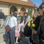 People line up outside a beige building with arches as adults hand yellow daffodils to children during a sunny daytime gathering.