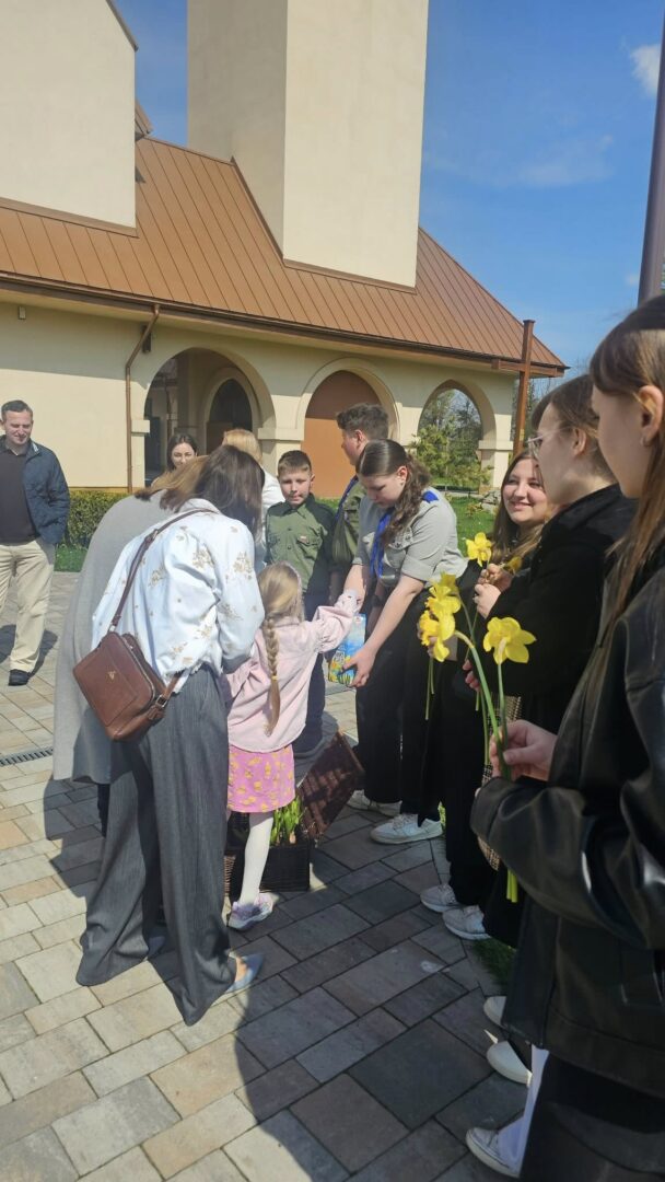 People line up outside a beige building with arches as adults hand yellow daffodils to children during a sunny daytime gathering.