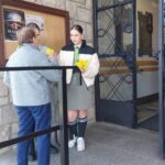 Young woman hands a bouquet of yellow flowers to an elderly woman at the doorway of a stone building.