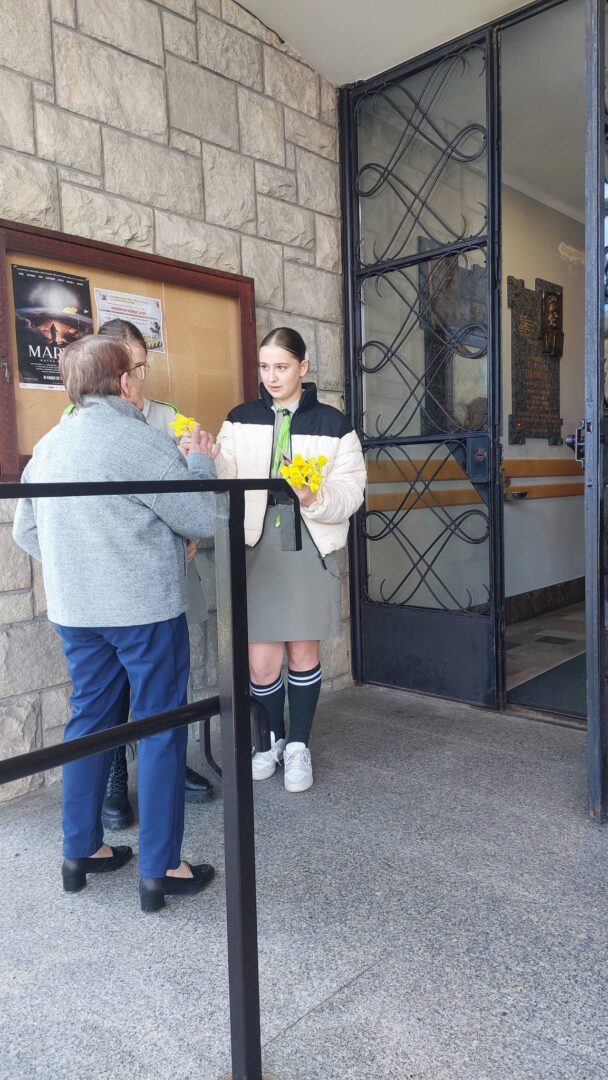 Young woman hands a bouquet of yellow flowers to an elderly woman at the doorway of a stone building.