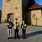 Three young men in smart jackets hold yellow daffodils and blue gift boxes outside a stone church on a sunny day.