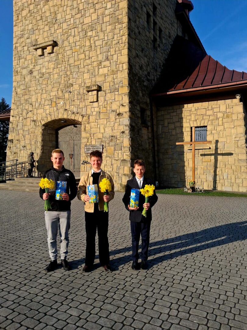 Three young men in smart jackets hold yellow daffodils and blue gift boxes outside a stone church on a sunny day.