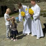 Two young men in white robes hand yellow daffodils to two children outdoors near a stone wall.