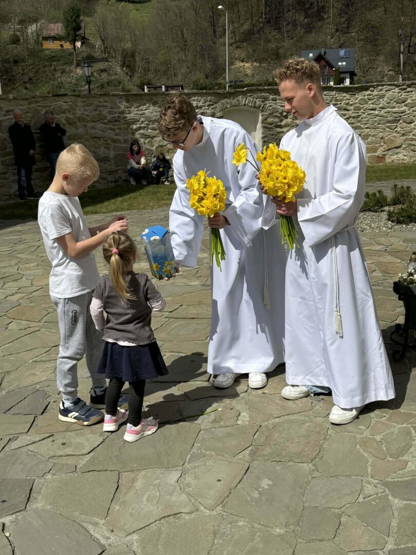 Two young men in white robes hand yellow daffodils to two children outdoors near a stone wall.
