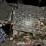 Nighttime view of a severely damaged house with a collapsed tiled roof, scattered rubble and exposed interior, including a metal stove vent.