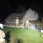 Nighttime scene of a collapsed stone house with firefighters inspecting rubble behind a safety barrier.