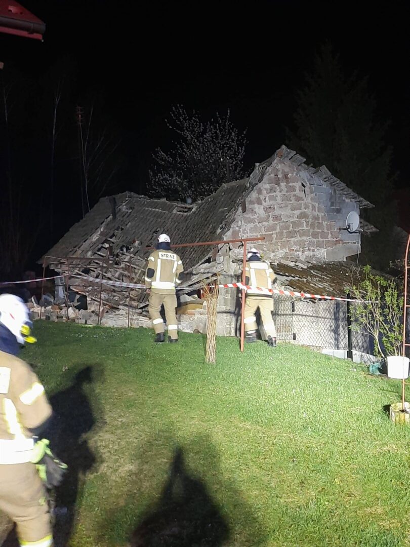 Nighttime scene of a collapsed stone house with firefighters inspecting rubble behind a safety barrier.