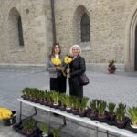 Two women stand by a table of potted daffodils outside a stone church, each holding yellow flowers and smiling.