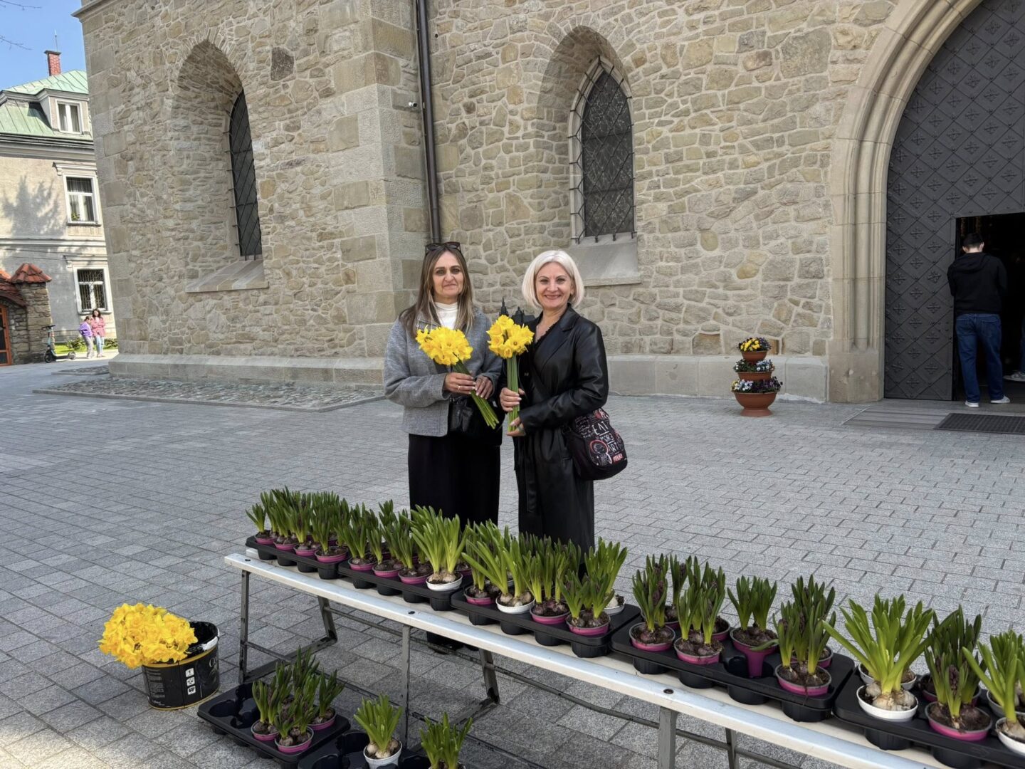 Two women stand by a table of potted daffodils outside a stone church, each holding yellow flowers and smiling.