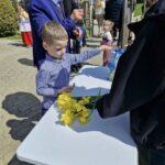 Young boy in a blue checkered shirt places yellow daffodils on a white folding table at an outdoor event, as an older man in a blue jacket smiles nearby and hands more flowers to him.