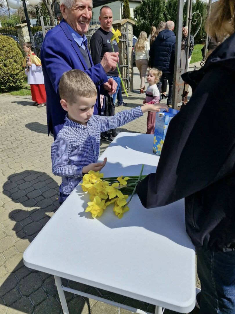 Young boy in a blue checkered shirt places yellow daffodils on a white folding table at an outdoor event, as an older man in a blue jacket smiles nearby and hands more flowers to him.