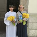 A young person in a white liturgical robe and a woman with glasses stand outside a doorway, each holding a bouquet of yellow daffodils; the woman also holds a blue gift tin with daffodil imagery and a card.