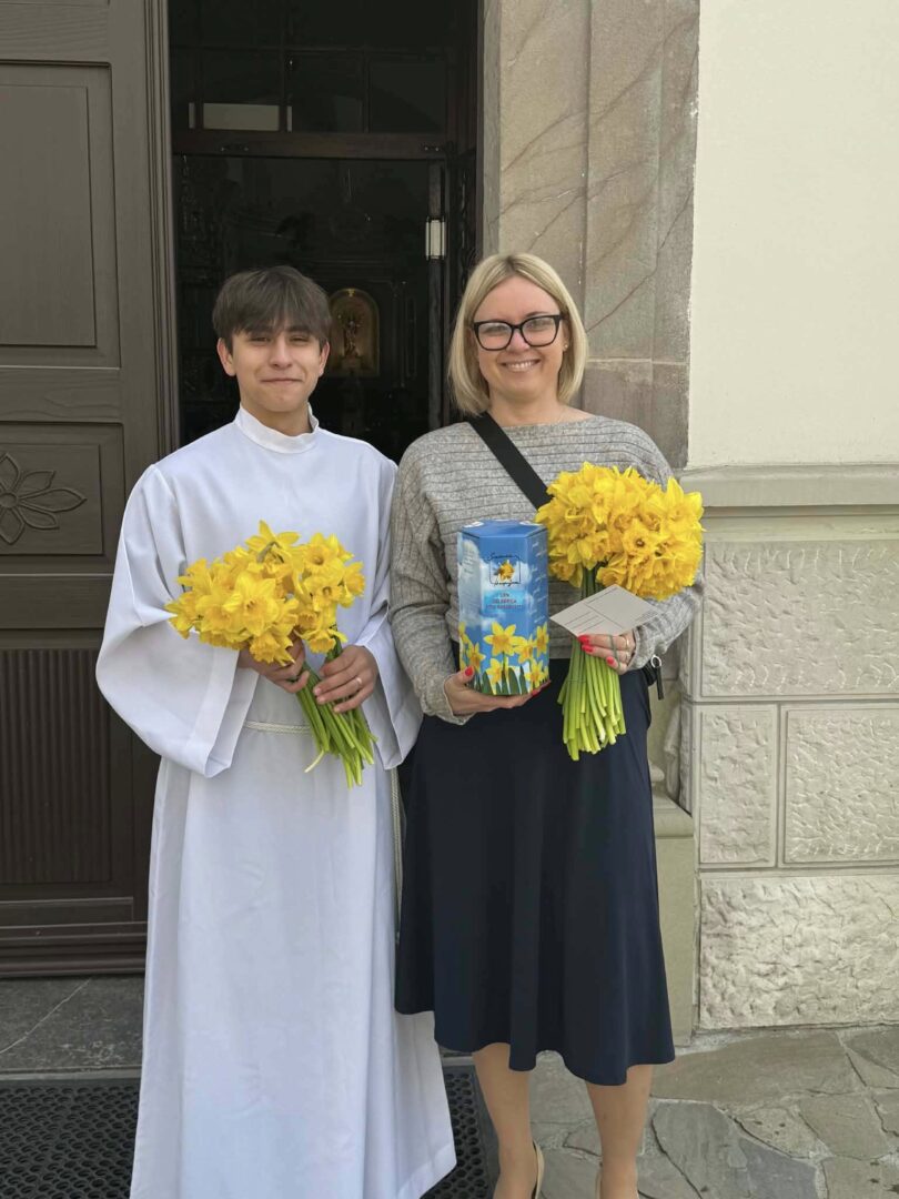 A young person in a white liturgical robe and a woman with glasses stand outside a doorway, each holding a bouquet of yellow daffodils; the woman also holds a blue gift tin with daffodil imagery and a card.