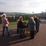 Group of people, mostly older adults, gather around a small wooden table with flowers outdoors on a paved plaza, hillside in the background on a sunny day.