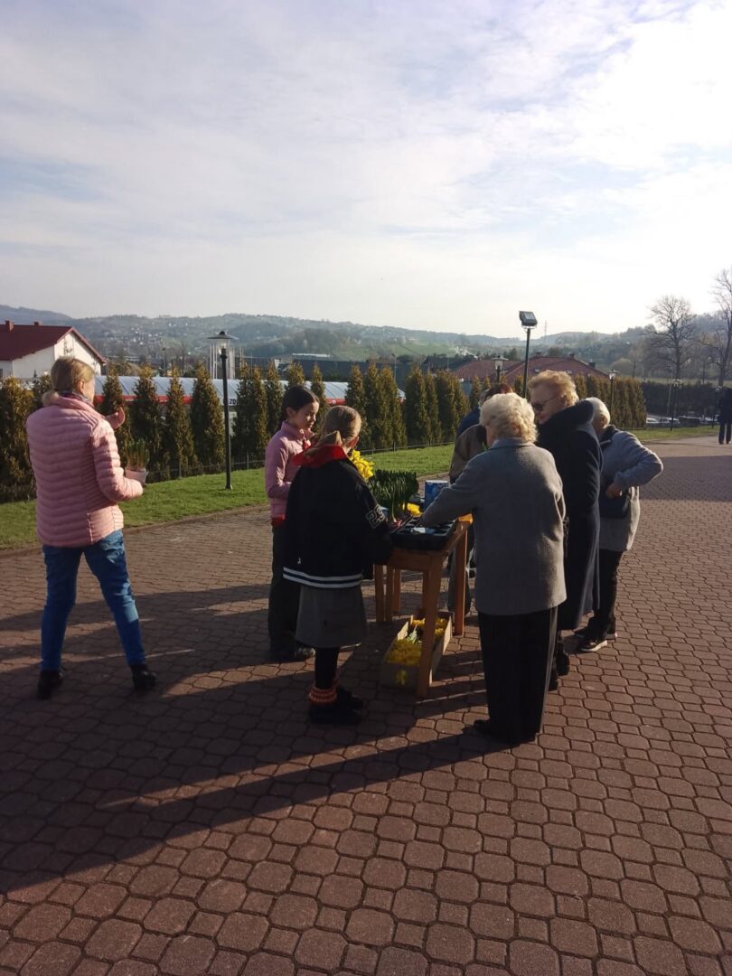 Group of people, mostly older adults, gather around a small wooden table with flowers outdoors on a paved plaza, hillside in the background on a sunny day.
