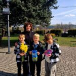 Woman with three boys outdoors, one holding yellow flowers, another a blue gift bag, the third looking at a pink item in his hands.