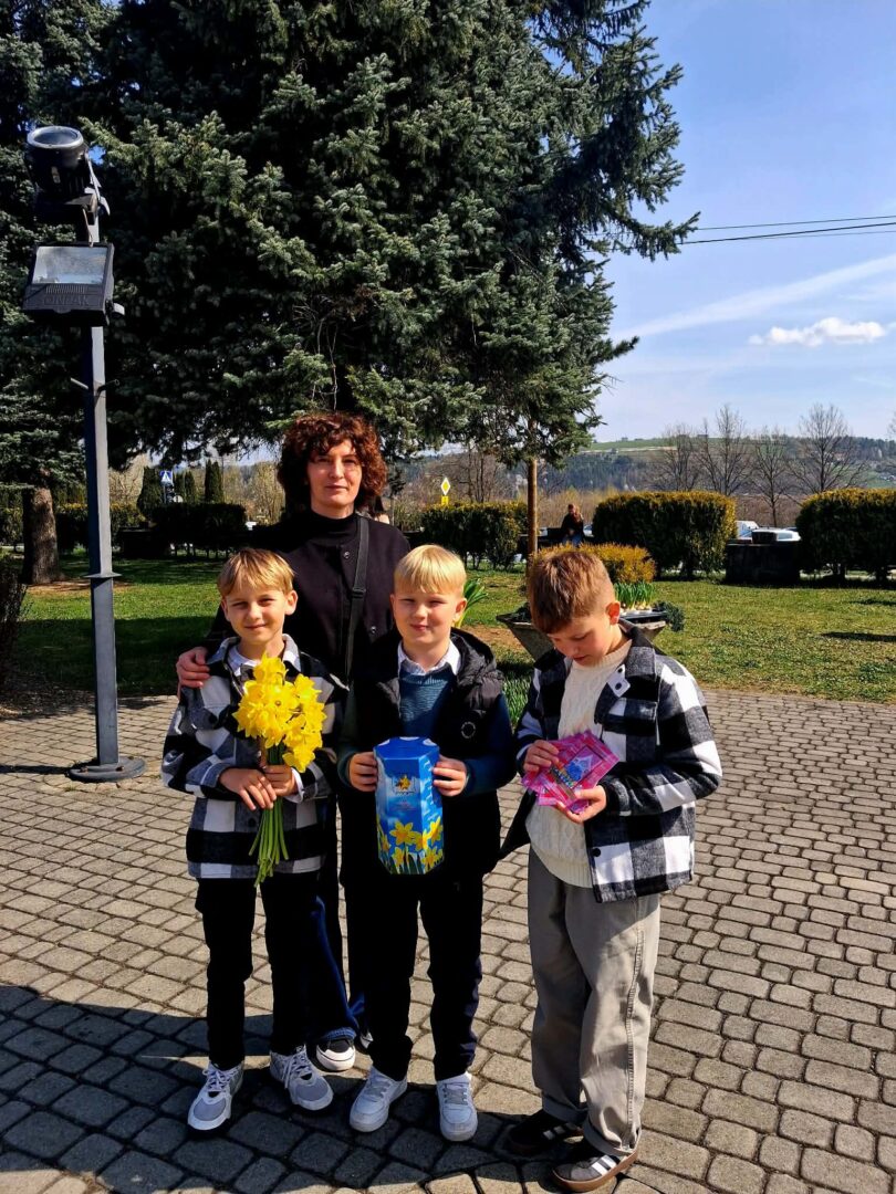 Woman with three boys outdoors, one holding yellow flowers, another a blue gift bag, the third looking at a pink item in his hands.