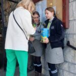 Adult woman speaks with two girl scouts outside a stone building, one girl holding a blue box.