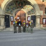 Five people in green uniforms stand in a decorative brick doorway, each holding yellow flowers and a blue book in front of a mosaic arch.