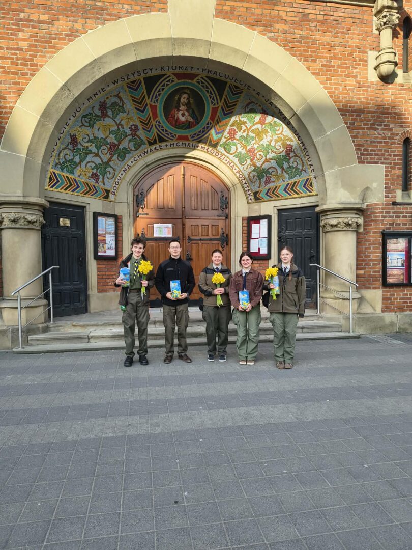 Five people in green uniforms stand in a decorative brick doorway, each holding yellow flowers and a blue book in front of a mosaic arch.