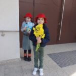 Two children wearing red hats stand outside a building; the front child holds a bright yellow bouquet toward the camera.