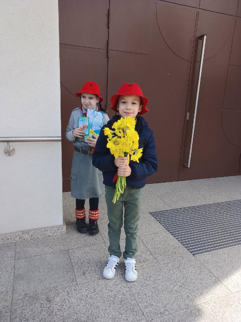 Two children wearing red hats stand outside a building; the front child holds a bright yellow bouquet toward the camera.