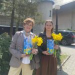 Two women stand outside, smiling, each holding a bouquet of yellow daffodils and a blue gift box with a QR code.