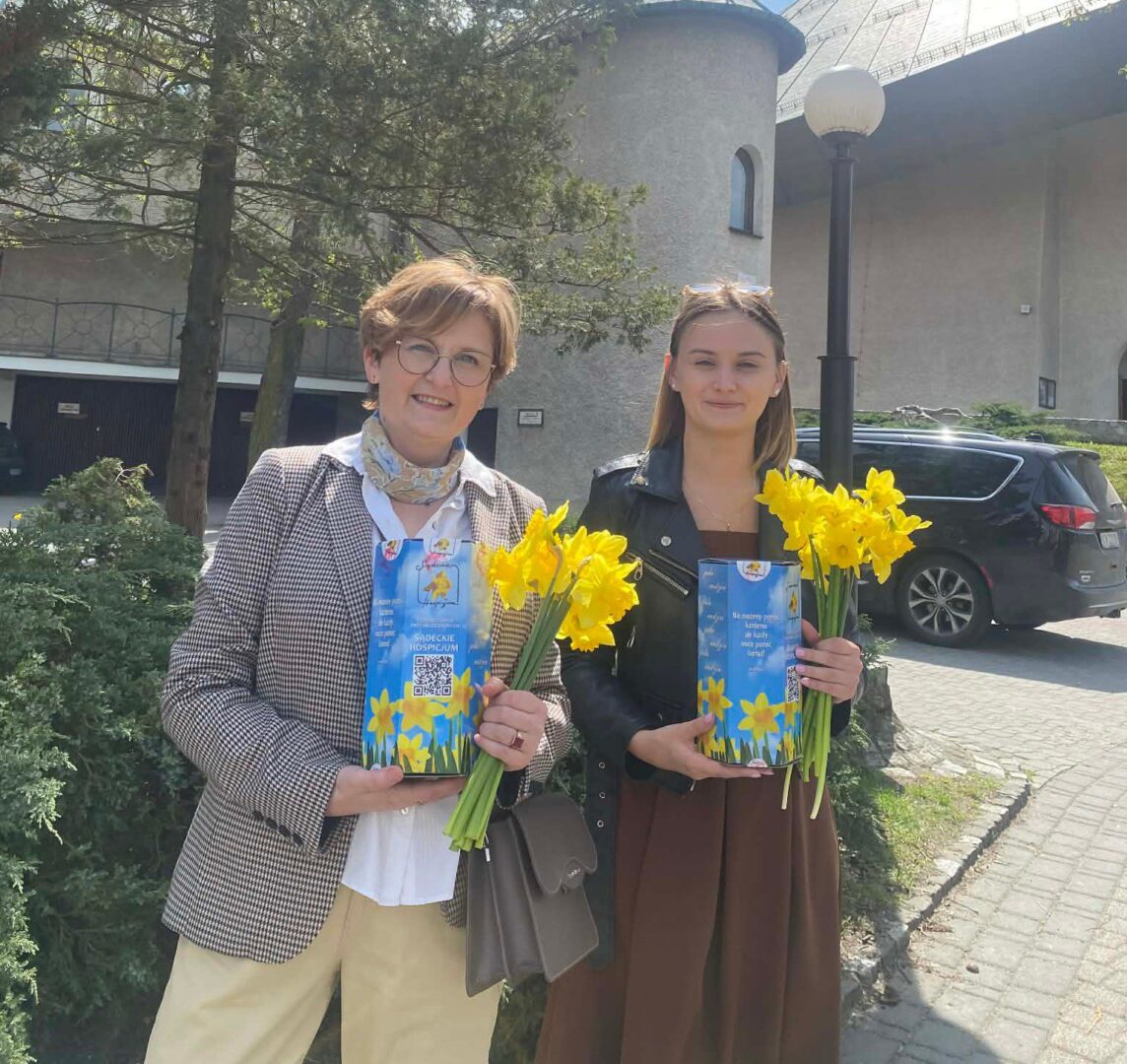 Two women stand outside, smiling, each holding a bouquet of yellow daffodils and a blue gift box with a QR code.