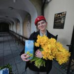 Young student in a red beret holds a bunch of bright yellow daffodils and a blue gift box outside a building with a plaque.