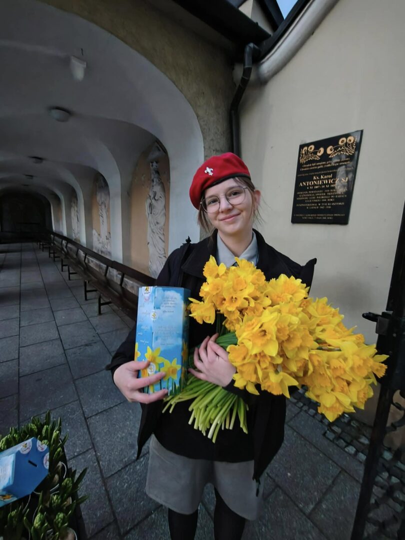 Young student in a red beret holds a bunch of bright yellow daffodils and a blue gift box outside a building with a plaque.