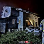 Firefighter in reflective gear stands near a partially collapsed stone house at night, rubble scattered in front.