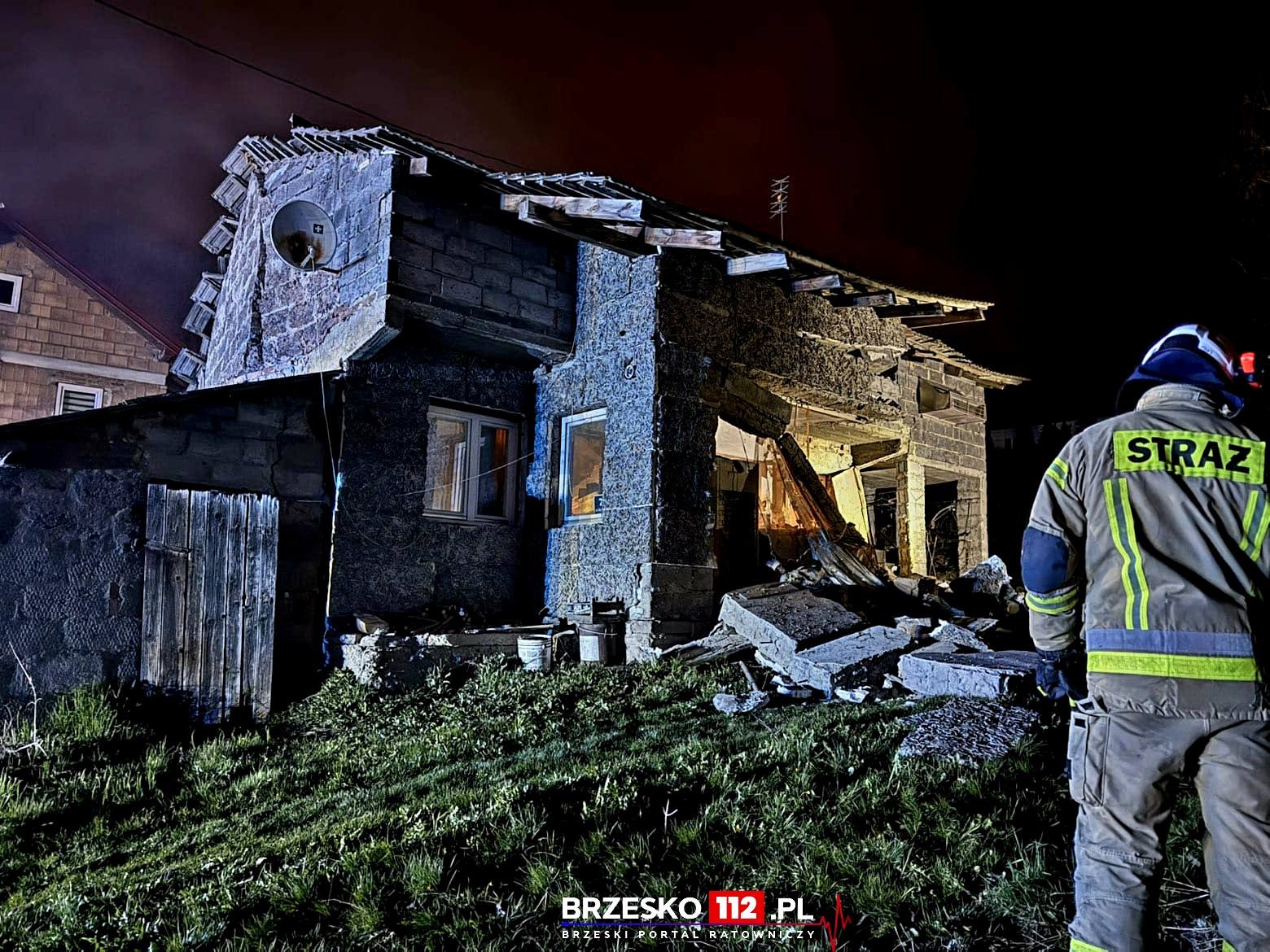Firefighter in reflective gear stands near a partially collapsed stone house at night, rubble scattered in front.