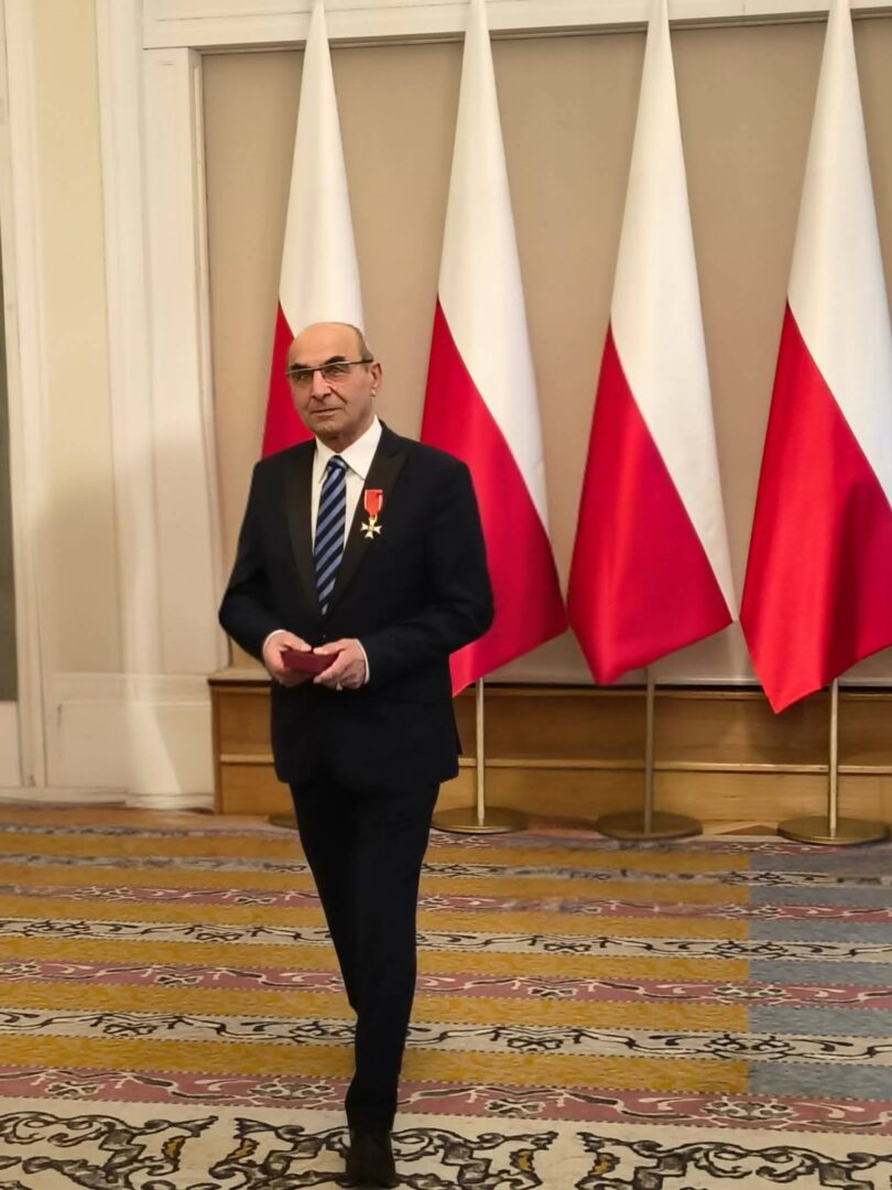 Man in a black suit and striped tie stands indoors, wearing a medal, with multiple Polish flags behind him and a patterned carpet underfoot.
