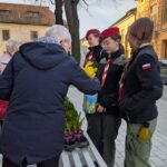 Elderly person giving yellow flowers to three young scouts in red berets on a city sidewalk; bench and potted plants nearby.