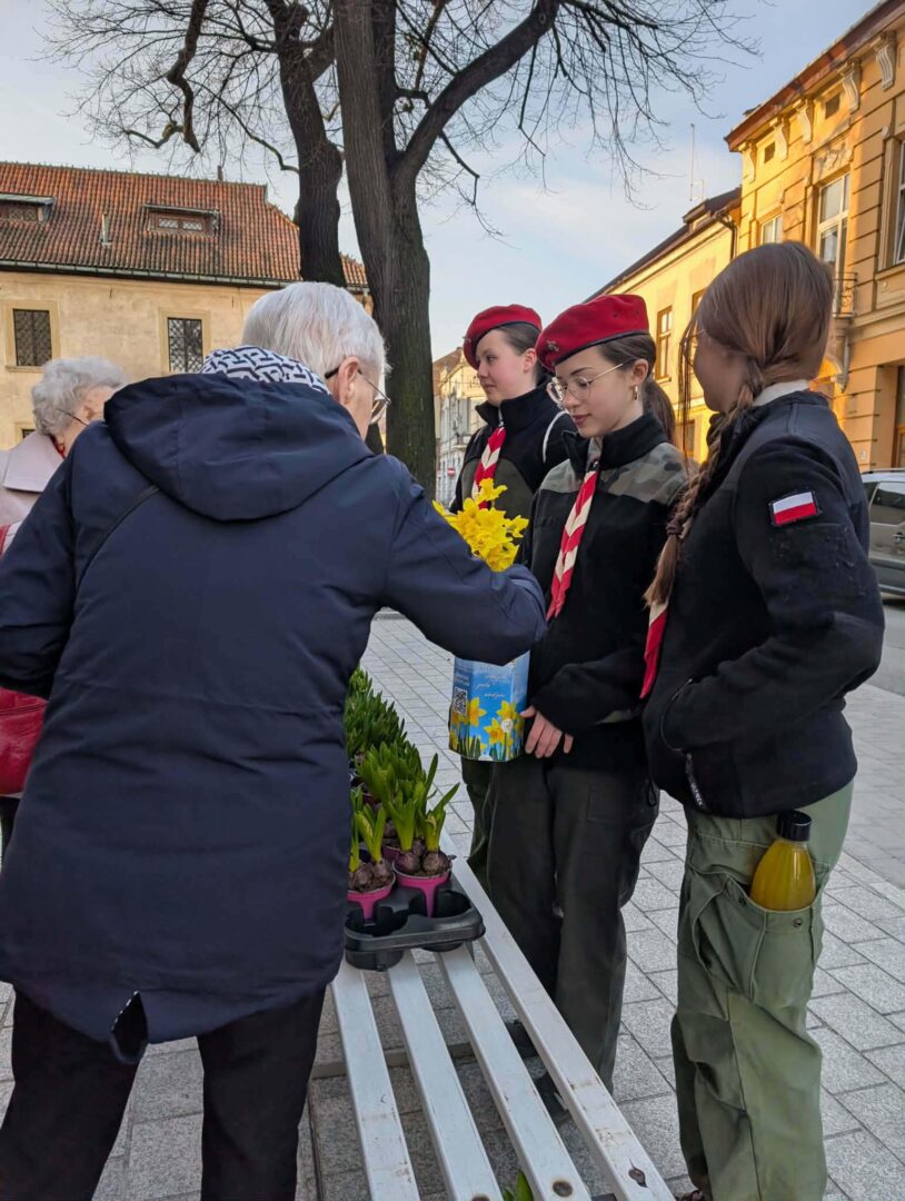 Elderly person giving yellow flowers to three young scouts in red berets on a city sidewalk; bench and potted plants nearby.