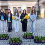 Five young women stand outside a building, holding yellow daffodil bouquets and plant boxes, with trays of potted plants on the ground in front of them.