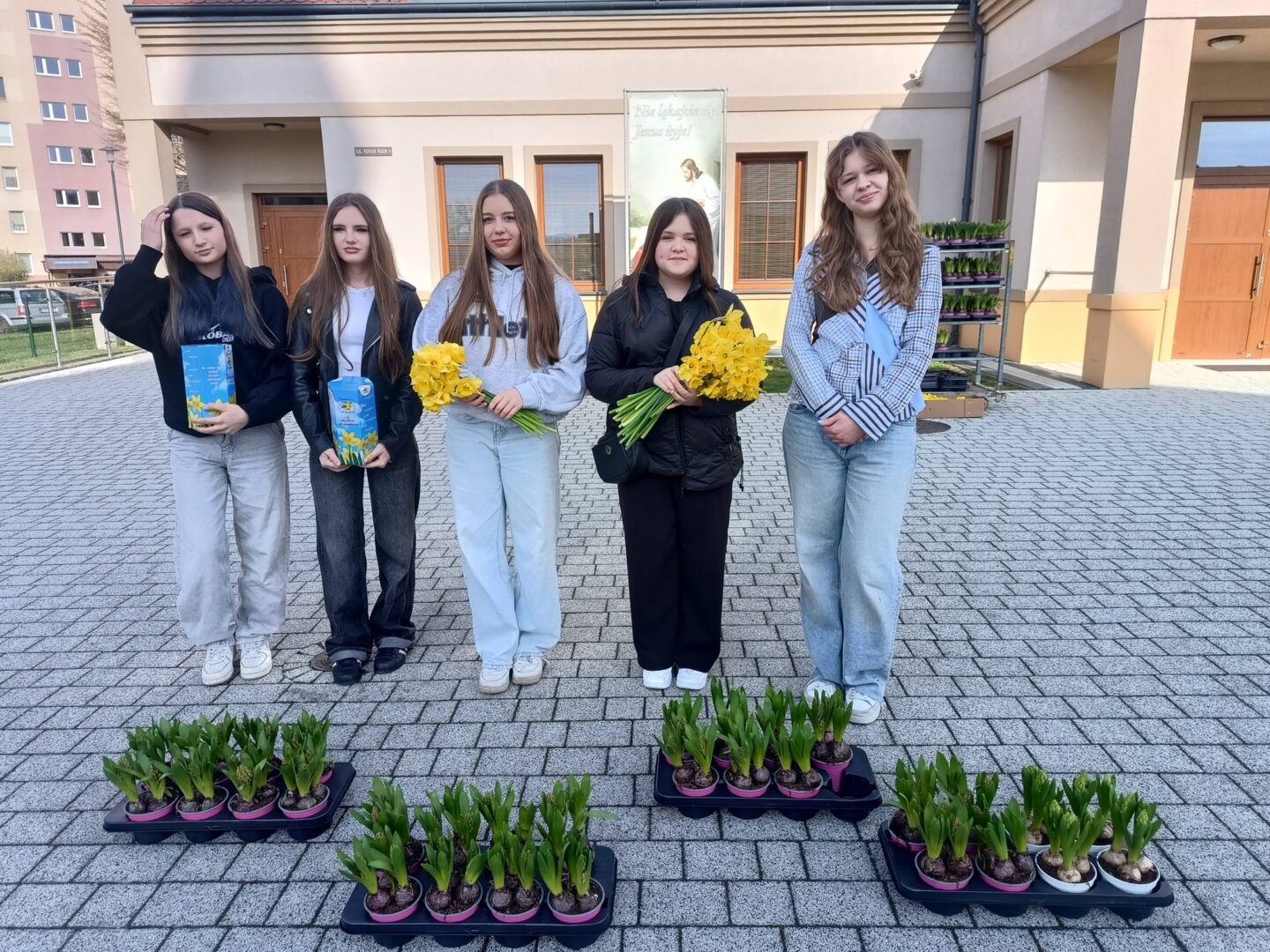Five young women stand outside a building, holding yellow daffodil bouquets and plant boxes, with trays of potted plants on the ground in front of them.
